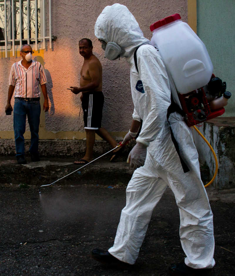 Worker in a protective suit with a spraying device on their back; in the background two people, only one wearing a face mask